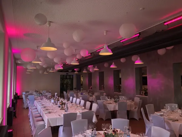 Saal mit langen Tafeln, Papierlampions und rosa Licht im Quartier77